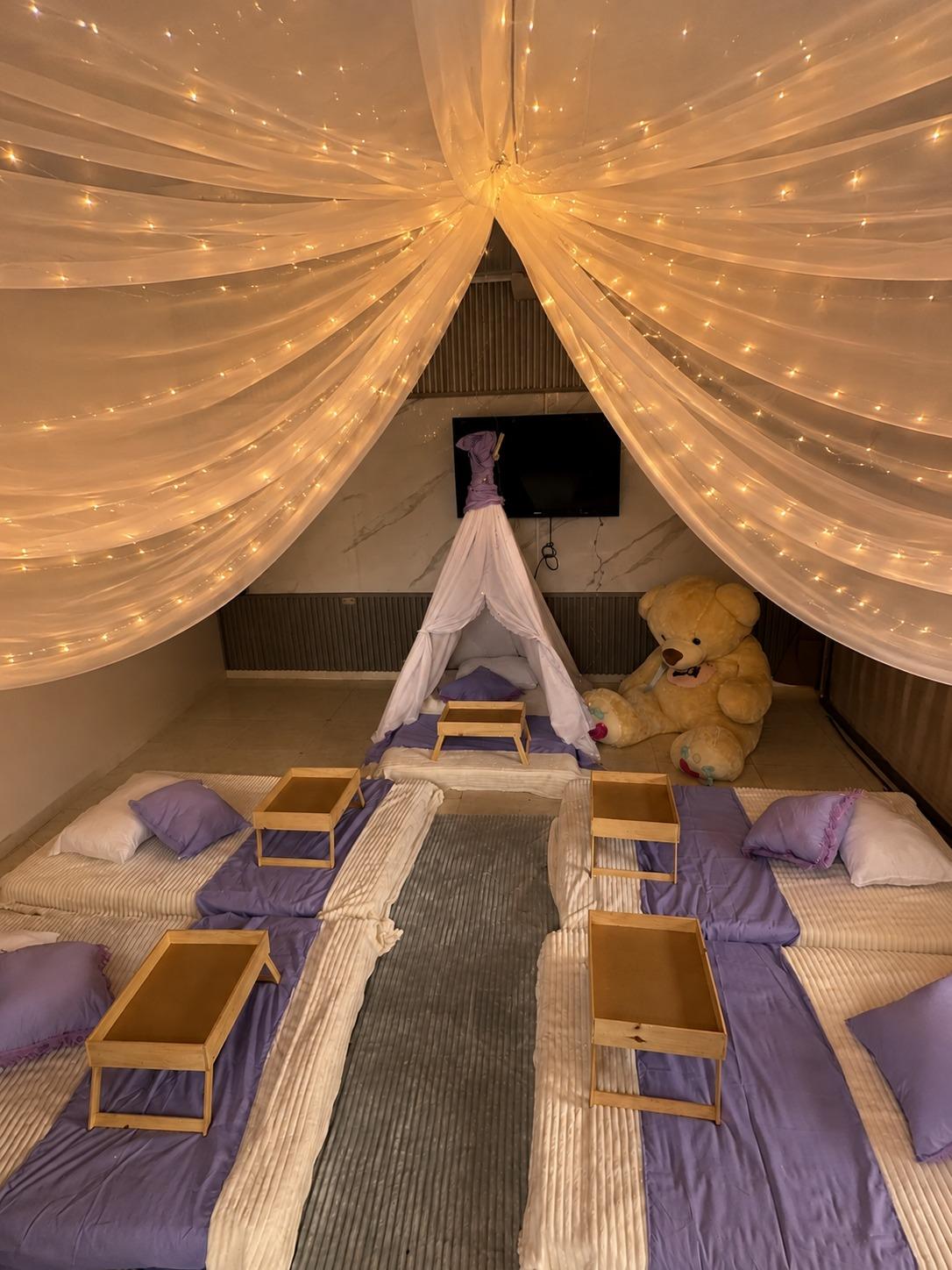Techo de velos con luces cálidas sobre una pijamada, con teepee, osito gigante y mesas desayunadoras