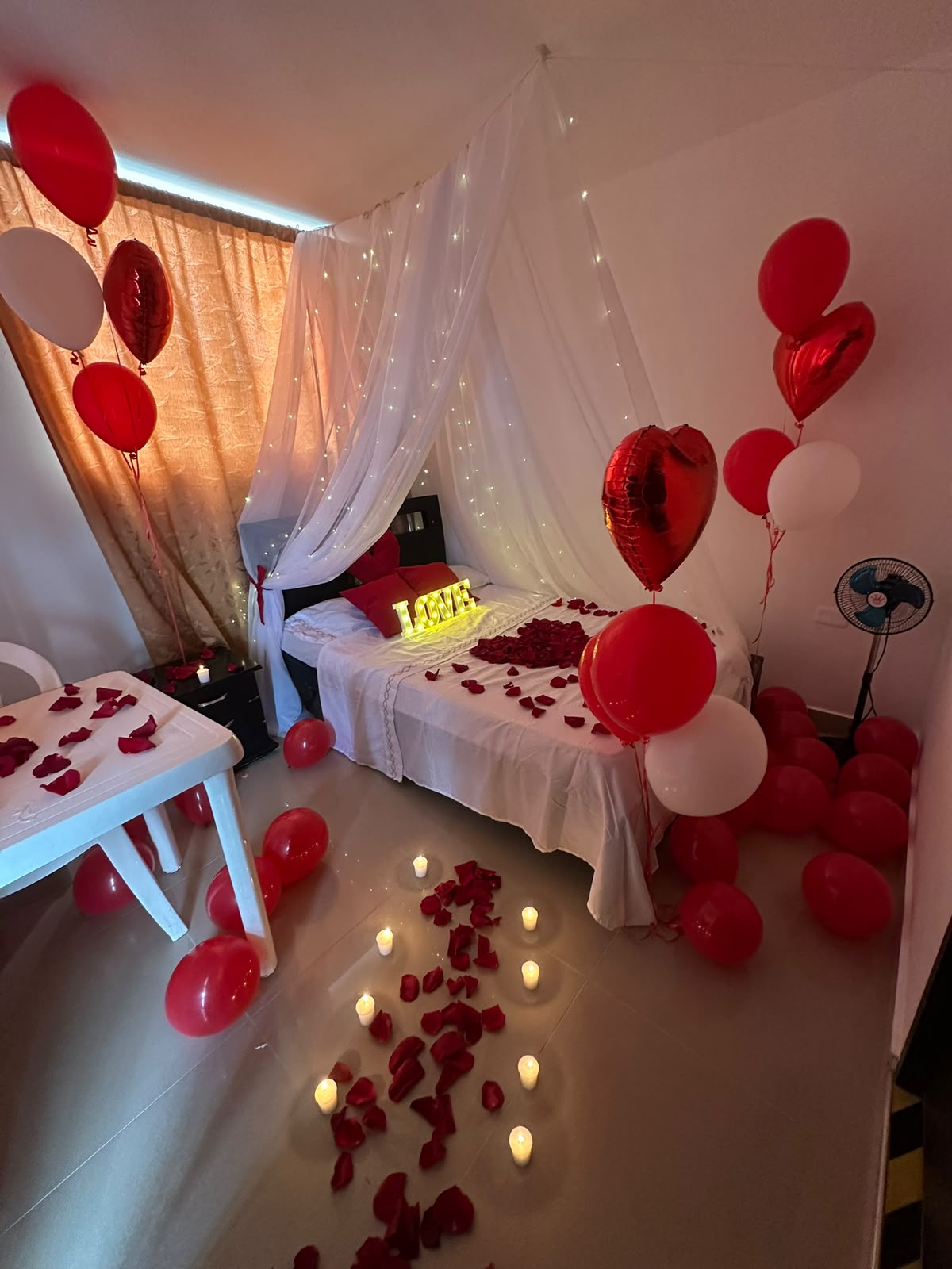 Decoración romántica de cuarto con velo de luces, pétalos de rosa y globos rojos, montada por Cony Decora